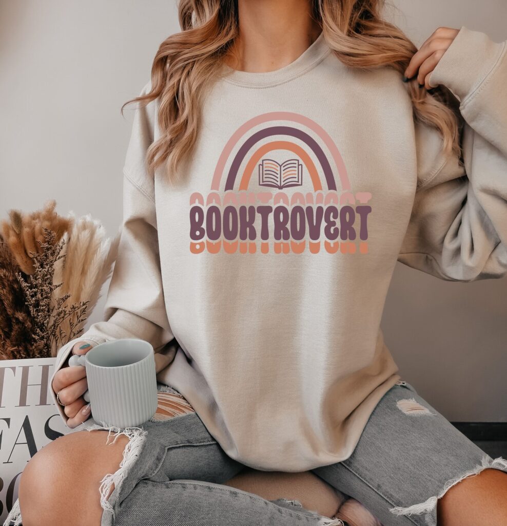 Woman wearing a sweatshirt decorated with the word BOOKTROVERT, an illustration of an open book and a rainbow above the word.