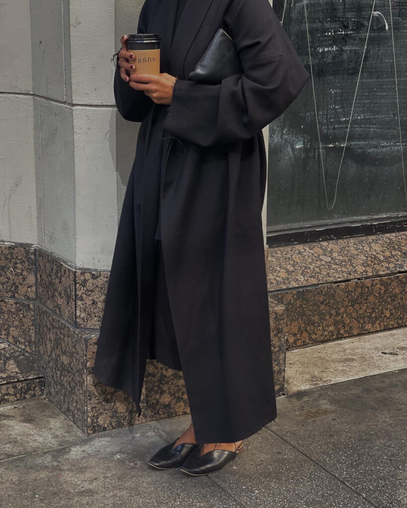 Photo of a woman in a minimalist black trench coat and unbranded leather flats suggesting a wardrobe inspired by quiet luxury.
